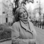 A stylish senior woman standing on a city sidewalk, smiling warmly in grayscale photography.